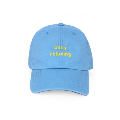 A sky blue baseball cap with the text 'busy relaxing' embroidered in yellow on the front.  Casual self-care inspired cap