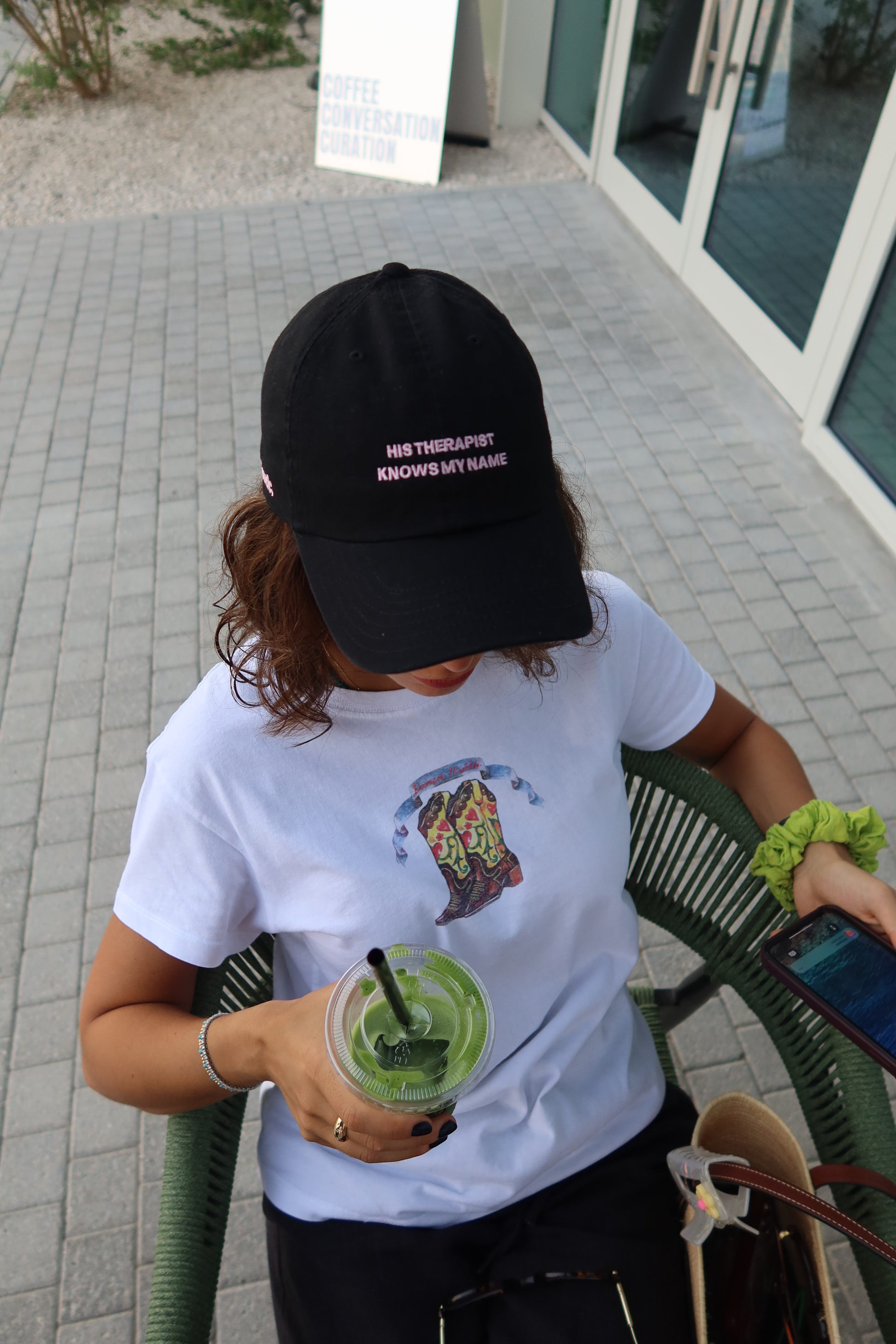 Person holding a green drink and a phone, wearing a black cap with text " his therapist knows my name' and white shirt with a colorful design.