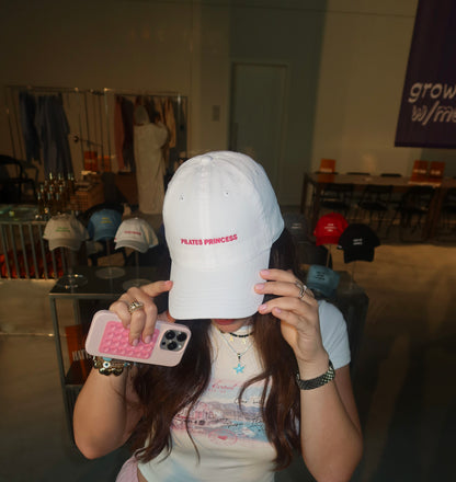 Person holding a white cap with 'Pilates Princess' text in a casual indoor setting.