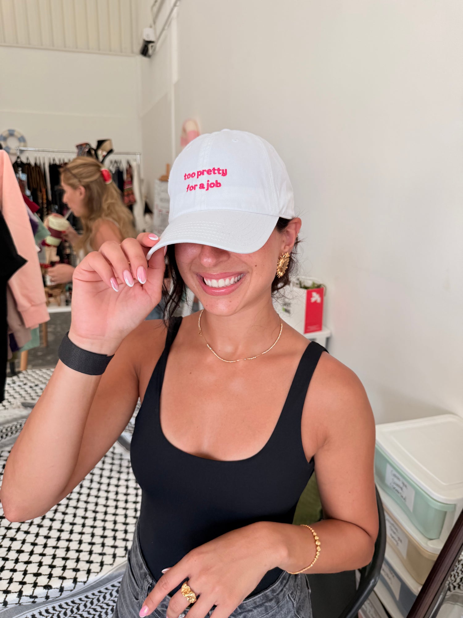 Woman wearing a white cap with text ' too pretty for a job', smiling indoors.