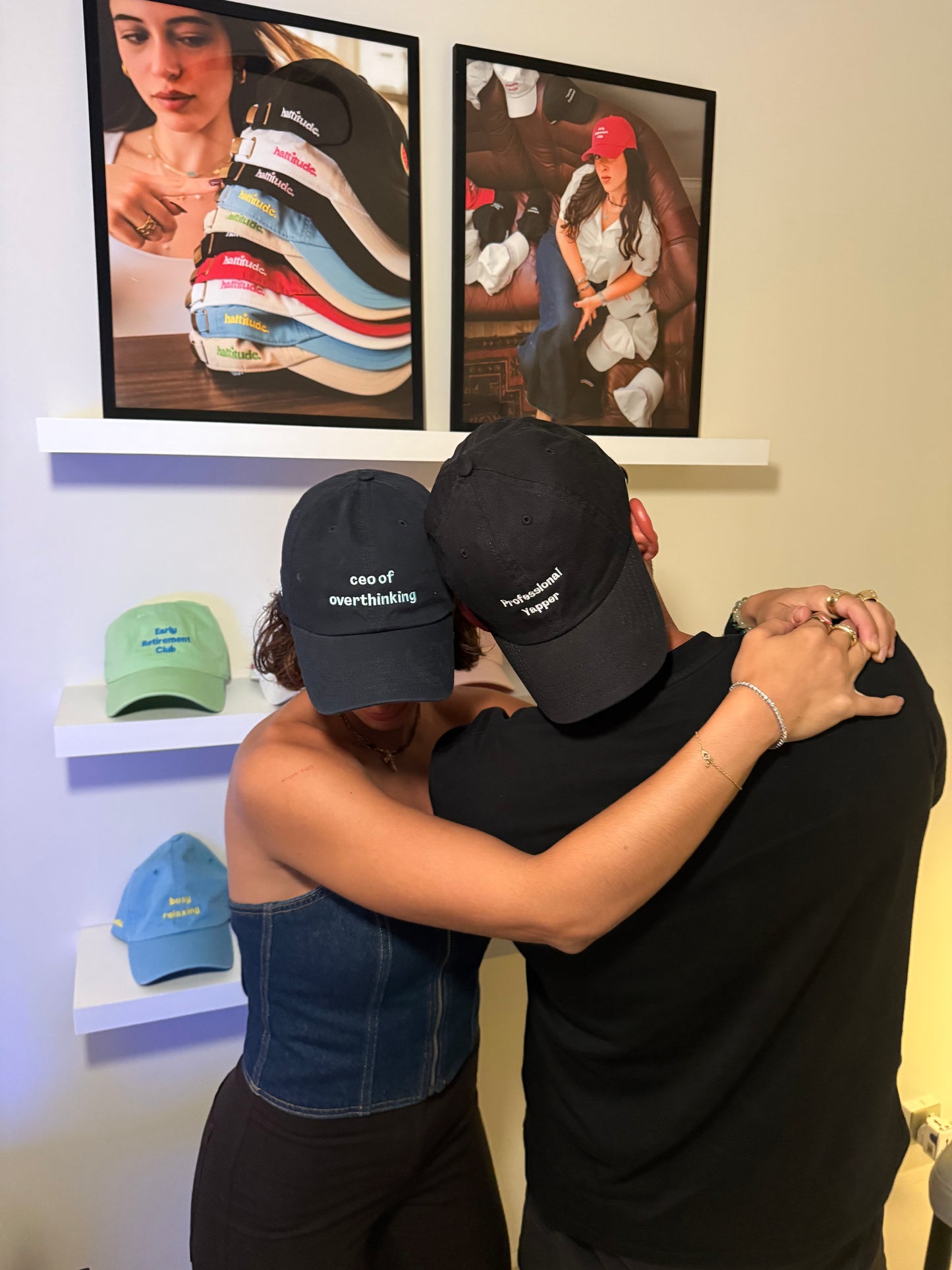 Two people wearing hattitude statement caps, standing in front of framed images on a wall.
